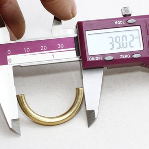 May include: A gold-colored, semi-circular metal object is measured with a digital caliper. The caliper displays a measurement of 39.02, likely millimeters. The object has a smooth, polished surface and appears to be a component or finding.