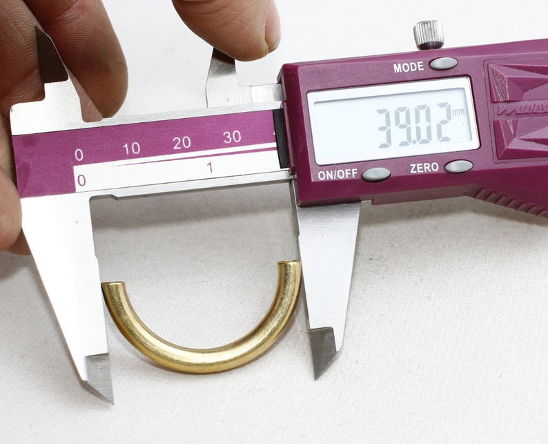 May include: A gold-colored, semi-circular metal object is measured with a digital caliper. The caliper displays a measurement of 39.02, likely millimeters. The object has a smooth, polished surface and appears to be a component or finding.