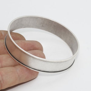 Könnte beinhalten: Ein silberfarbenes Armreif-Armband mit einem flachen, breiten Band und einer gebürsteten Metalloberfläche. Das Armband hat ein schlichtes, modernes Design und wird vor einem weißen Hintergrund gezeigt. Die Breite des Armbands beträgt ca. 2,5 cm.
