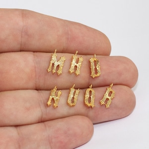 Puede incluir: Primer plano de varios peque&ntilde;os colgantes con forma de letras doradas. Las letras est&aacute;n estilizadas con un dise&ntilde;o de rayos de sol y cada una tiene un peque&ntilde;o lazo en la parte superior para sujetarlas. Las letras se sostienen en una mano.