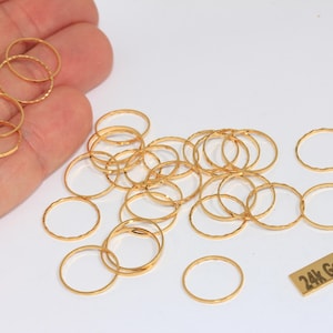 Puede incluir: Una colección de pequeños anillos dorados, algunos con superficies texturizadas, se encuentran sobre un fondo blanco. También hay una pequeña etiqueta rectangular con el texto "24k Gold Plated". Los anillos varían en tamaño y diseño.