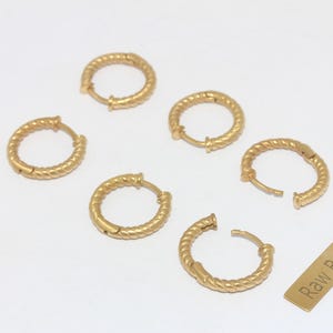 Puede incluir: Seis pendientes de aro dorados con un diseño de cuerda trenzada se muestran sobre una superficie blanca. Los pendientes están abiertos y cerrados, mostrando el mecanismo de cierre. También hay una pequeña etiqueta rectangular con el texto "Raw Brass".