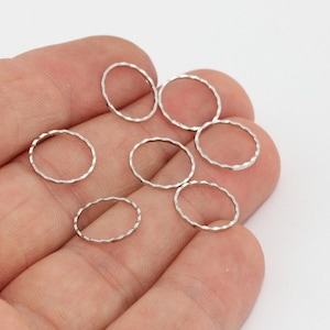 May include: A collection of small, silver-colored, textured oval-shaped rings. The rings are held in a person's hand against a white background. These jewelry findings are ideal for crafting earrings, necklaces, or other accessories.