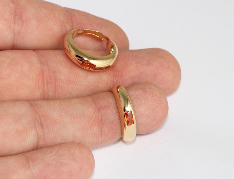 May include: Two gold-colored rings with a rectangular orange gemstone. One ring is on a finger, and the other is lying flat. The rings have a smooth, polished finish and a simple, elegant design. Jewelry.