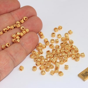 Puede incluir: Una colección de pequeñas cuentas cilíndricas chapadas en oro mate con una superficie texturizada y acanalada. Una etiqueta metálica rectangular con el texto "Matt Gold Plated" se sitúa junto a las cuentas sueltas sostenidas en una palma.