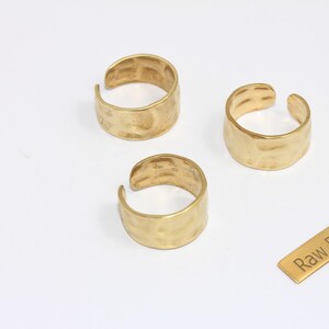 May include: Three gold-colored, hammered-texture rings with an open design are displayed on a white surface. A small rectangular tag with the text "Raw Brass" is also present. The rings are likely for jewelry.