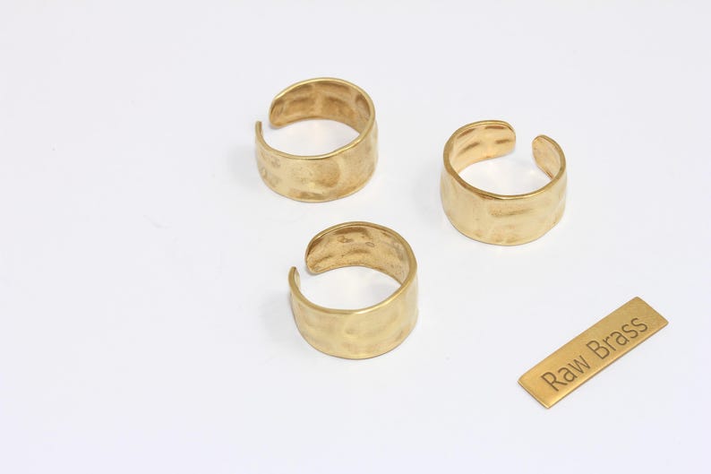 May include: Three gold-colored, hammered-texture rings with an open design are displayed on a white surface. A small rectangular tag with the text "Raw Brass" is also present. The rings are likely for jewelry.