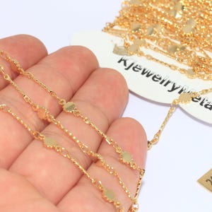Pode incluir: Close-up de joias de corrente banhadas a ouro. A corrente apresenta pequenos pingentes planos em forma de flor e elos texturizados. Uma etiqueta diz "kjewelrymetal" e uma pequena etiqueta dourada diz "24k Gold Plated".