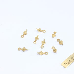Könnte beinhalten: Eine Sammlung kleiner, goldfarbener Messinganhänger mit dekorativem Design, verstreut auf einer weißen Oberfläche. Die Anhänger haben an einem Ende eine Schlaufe und am anderen eine detaillierte Form. Ein rechteckiges Etikett mit dem Text "Raw Brass" ist ebenfalls vorhanden.