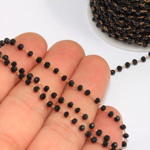 Puede incluir: Primer plano de un hilo de cuentas negras facetadas sobre una cadena dorada. Las cuentas son pequeñas y están espaciadas uniformemente. Un carrete de la misma cadena de cuentas es visible en el fondo.