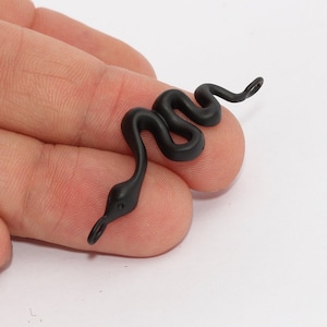 Peut inclure: Un charm en forme de serpent noir mat avec un corps incurvé et une petite tête arrondie. Le charme est tenu dans une main sur fond blanc. Le serpent a une petite boucle au bout de sa queue et une petite boucle au bout de sa tête.