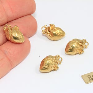 May include: Four small, gold-coloured heart charms with detailed anatomical features. One charm is held in a hand, the others are on a white surface. A small rectangular tag reads "Raw Brass".
