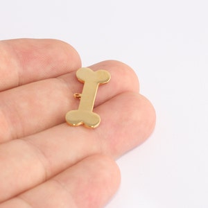 Puede incluir: Un dije dorado en forma de hueso con un pequeño lazo en la parte superior para sujetarlo a un collar o pulsera. El dije tiene un acabado pulido y está sobre un fondo blanco. Este dije es adecuado para los amantes de las mascotas.