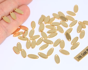 May include: A collection of small, gold-colored, oval-shaped charms with a hole at the top. The charms are scattered on a white surface, with some held in a hand. A small rectangular tag reads "24k Gold Plated".