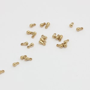 Puede incluir: Pequeños abalorios dorados en forma de lágrima con un lazo en la parte superior están dispersos sobre una superficie blanca. Estos abalorios son probablemente para la fabricación de joyas u otros proyectos de artesanía. Acabado metálico liso.