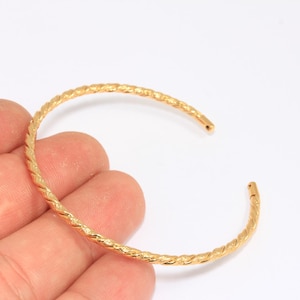 May include: A gold-colored open cuff bracelet with a textured, twisted rope design. The bracelet is held against a white background, showcasing its intricate details and warm metallic hue. This piece of jewelry is a stylish accessory.