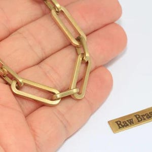 Puede incluir: Una cadena dorada con eslabones rectangulares se sostiene en una mano. Una pequeña etiqueta rectangular con las palabras "Raw Brass" está al lado de la cadena. La cadena y la etiqueta están sobre un fondo blanco.