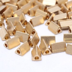 May include: A collection of small, gold-colored metal beads. Some beads are rectangular, while others are triangular with a hole at one end. The beads are scattered on a white surface.