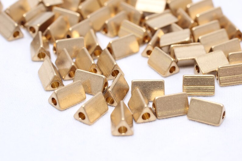 May include: A collection of small, gold-colored metal beads. Some beads are rectangular, while others are triangular with a hole at one end. The beads are scattered on a white surface.