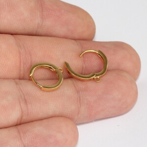 May include: Two gold-colored hoop earrings. Each earring is a small, rounded hoop with a hinged closure. The earrings are shown against a neutral background, highlighting their simple, elegant design. The earrings are made of a shiny, metallic material.