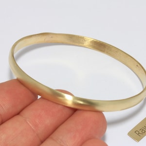 Puede incluir: Un brazalete de latón de color dorado. El brazalete tiene un diseño simple y redondeado. Una pequeña etiqueta de latón con las palabras "Raw Brass" está al lado del brazalete.
