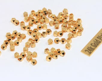Puede incluir: Un conjunto de pequeñas cuentas doradas facetadas dispersas sobre una superficie blanca. También hay una pequeña etiqueta rectangular con el texto "24k Gold Plated". Adecuado para la creación de joyas.