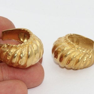 Può includere: Due orecchini a cerchio color oro con un design strutturato e scanalato. Gli orecchini hanno una forma arrotondata con una superficie strutturata e un retro liscio e aperto. Presentati su sfondo bianco.