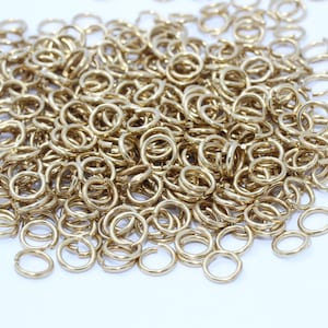 May include: A pile of small, gold-colored jump rings. These open rings are round and appear to be made of metal. The rings are scattered across a white surface, with some rings overlapping.