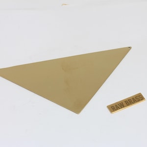 Peut inclure: Un triangle en laiton brut avec deux petits trous et une étiquette rectangulaire portant l'inscription "RAW BRASS". Le laiton a une teinte dorée et est posé sur un fond blanc.