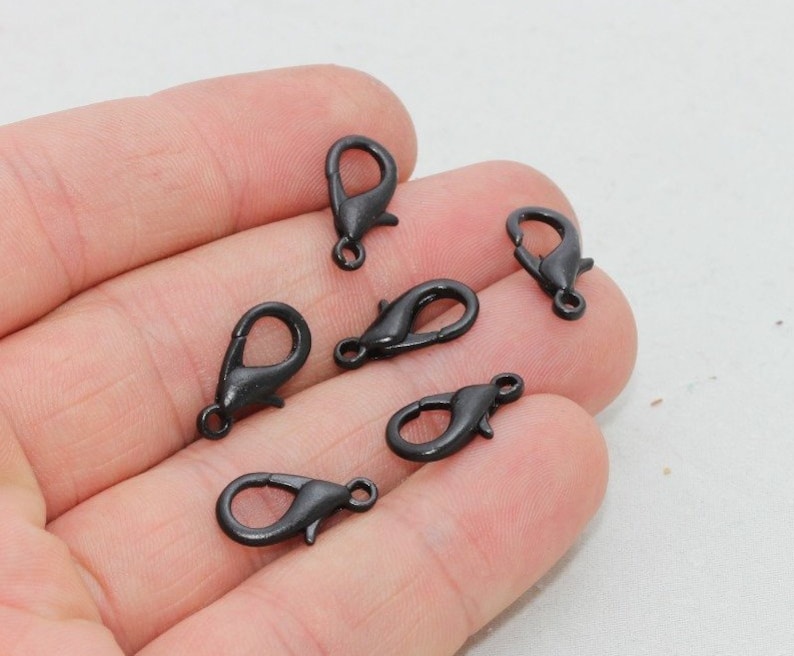 May include: Six small, black metal lobster claw clasps are displayed. Each clasp features a spring-loaded lever and a small loop for attaching to jewellery or other items. The clasps are shown against a neutral background.