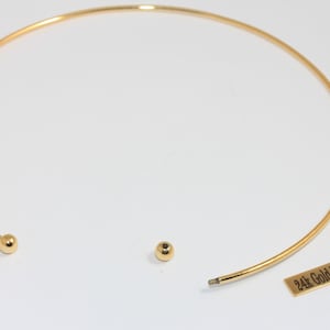 Pode incluir: Um colar gargantilha banhado a ouro com um design simples e elegante. O colar é uma faixa fina e curva com uma pequena bola em cada extremidade. Uma pequena bola separada e uma etiqueta com a inscrição "24k Gold Plated" também são visíveis.