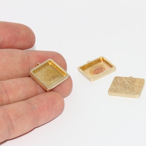 Può includere: Primo piano di tre castoni quadrati dorati per gioielli. Uno è tenuto in mano, mostrando la sua superficie strutturata e un piccolo anello per attaccare una catena. Gli altri due castoni sono su una superficie bianca.