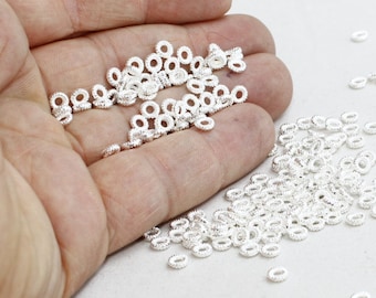 May include: A collection of small, silver-colored metal ring beads with a textured surface. The beads are scattered across a hand, with some clustered together and others spread out. These jewelry-making components are ideal for crafting.
