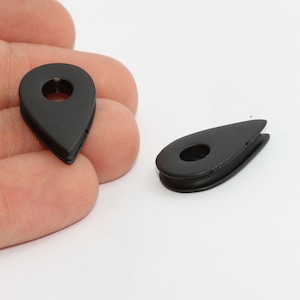 May include: Two black, teardrop-shaped metal grommets with a central hole. The grommets have a matte finish and are designed for use in various crafting and DIY projects. One grommet is held in a hand, while the other is lying flat.