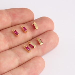以下が含まれることがあります： 中央に明るいピンク色の長方形の宝石がセットされた、小さな金色の長方形のチャーム。チャームの上部には、ジュエリーに取り付けるための小さなループが付いています。いくつかのチャームが明るい背景に散らばっています。
