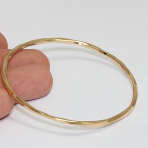 Puede incluir: Un brazalete de color dorado con una textura martillada. El brazalete circular se muestra sobre un fondo blanco. El brazalete es una pieza de joyería sencilla y elegante.