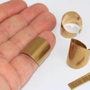 Peut inclure: Trois bagues larges ouvertes en laiton brut avec une finition métallique lisse. Une bague est placée sur un doigt, tandis que deux autres sont posées à côté avec une petite plaque rectangulaire en laiton portant l'inscription RAW BRASS.