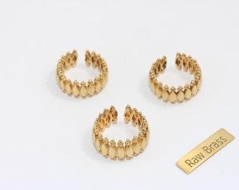 May include: Three gold-colored adjustable rings with a repeating teardrop design are displayed on a white surface. A small, rectangular gold tag with the words "Raw Brass" is also present. The rings are open-ended.