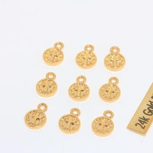 K&ouml;nnte beinhalten: Eine Sammlung goldfarbener, runder Anh&auml;nger mit Kreuzdesign, jeweils mit einer kleinen Schlaufe oben. Ein rechteckiger Anh&auml;nger tr&auml;gt die Aufschrift "24k Gold Plated". Diese Anh&auml;nger sind wahrscheinlich f&uuml;r die Schmuckherstellung oder das Basteln gedacht.
