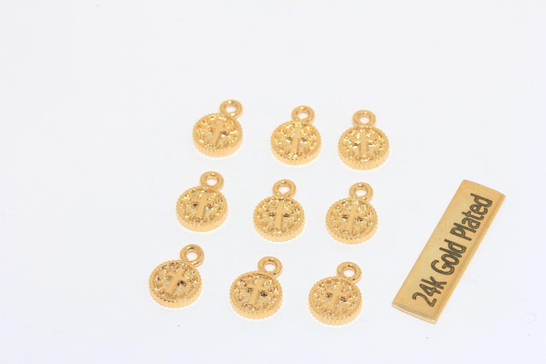 K&ouml;nnte beinhalten: Eine Sammlung goldfarbener, runder Anh&auml;nger mit Kreuzdesign, jeweils mit einer kleinen Schlaufe oben. Ein rechteckiger Anh&auml;nger tr&auml;gt die Aufschrift "24k Gold Plated". Diese Anh&auml;nger sind wahrscheinlich f&uuml;r die Schmuckherstellung oder das Basteln gedacht.