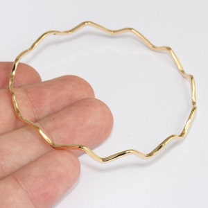 Può includere: Un braccialetto a cerchio ondulato color oro. Il braccialetto ha una forma irregolare e organica, con una serie di curve e angoli. Il metallo ha una finitura lucida, che riflette la luce. Il braccialetto è mostrato su uno sfondo bianco.