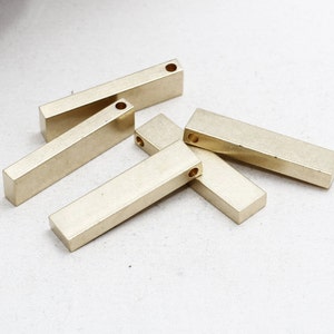 Può includere: Diverse barre rettangolari in metallo color oro, ciascuna con un piccolo foro praticato a un'estremità. Le barre sono disposte su una superficie bianca. Questi componenti sono probabilmente destinati alla creazione di gioielli o ad altri progetti artigianali.