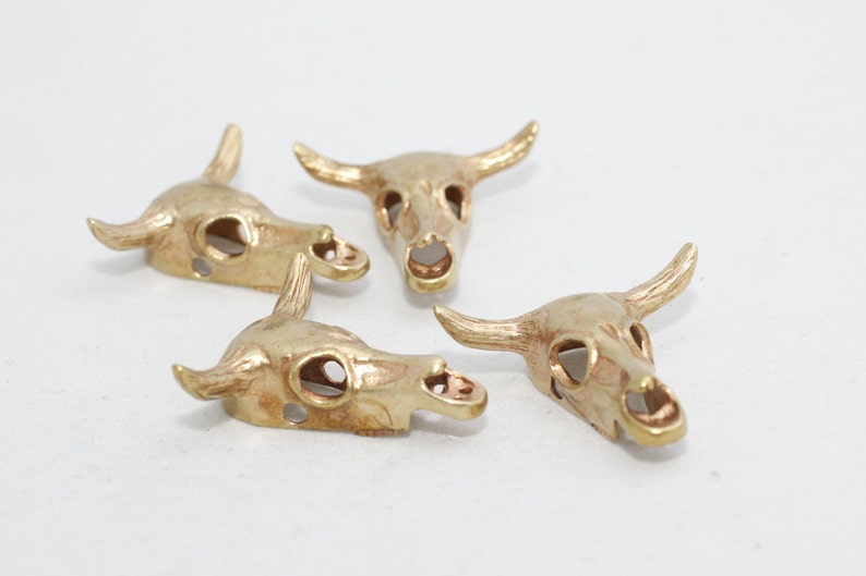 May include: Four small, gold-colored bull skull charms with horns and eye sockets are arranged on a white surface. The skulls are detailed with open eye sockets and nasal cavities, and are likely for jewelry making.