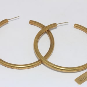 Peut inclure: Une paire de créoles dorées en laiton brut. Les boucles d'oreilles sont circulaires et présentent un design simple et minimaliste. Une petite étiquette rectangulaire avec les mots "Raw Brass" est également visible.