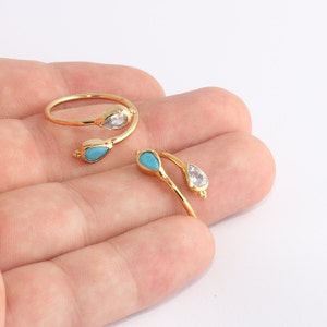 Puede incluir: Dos anillos dorados con detalles en forma de lágrima. Un anillo presenta una piedra transparente y una piedra turquesa, el otro una turquesa y una piedra transparente. Los anillos se muestran sobre un fondo claro.