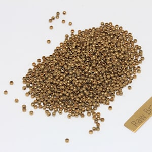 Può includere: Un mucchio di piccole perline dorate sparse su una superficie bianca. È presente anche una piccola etichetta in ottone con le parole "Raw Brass". Queste perline sono probabilmente utilizzate per la creazione di gioielli o altri progetti artigianali.