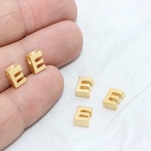 May include: Gold-colored metal letter "E" beads. The beads are cube-shaped with the letter cut out. Two beads are held in a hand, and three are scattered on a white surface. These small beads are ideal for jewelry making.