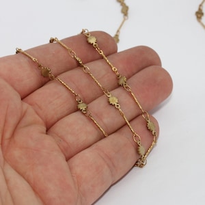 May include: Close-up of a delicate gold-colored necklace with small, flower-shaped charms. The necklace is composed of thin, twisted chain links and small connecting rings. The background is a plain white surface.