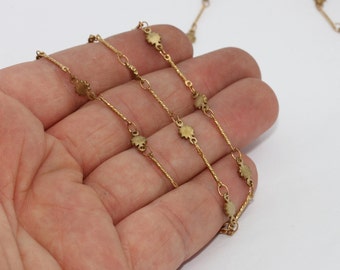 May include: Close-up of a delicate gold-colored necklace with small, flower-shaped charms. The necklace is composed of thin, twisted chain links and small connecting rings. The background is a plain white surface.