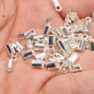 May include: A pile of small, silver-coloured crimp end findings for jewellery making. Each finding has a rectangular body with a loop at one end and a flat tab at the other, designed to secure cords or ribbons.